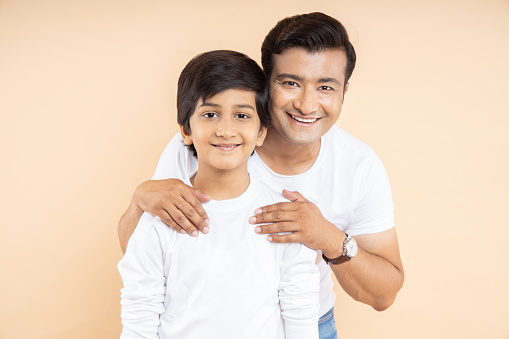 Happy young indian father with his son wearing white casual t-shirt standing over isolated beige background.