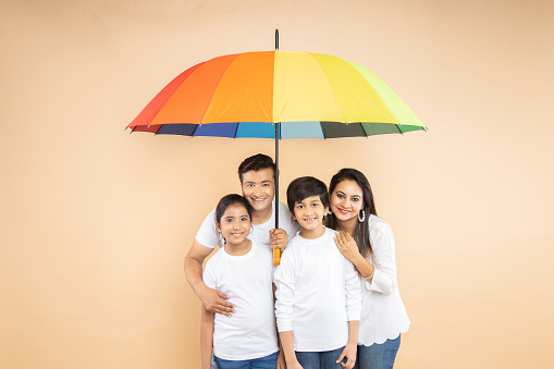 Happy indian family standing under big multicolor or colorful umbrella isolated on beige background. parents and children. Life and health insurance Safety concept.