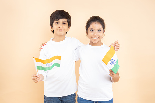 Cheerful indian little boy and girls kids wearing white t-shirt holding indian national flag isolated on beige background, happy independence day, 15th of august, republic day, Protestantism concept.