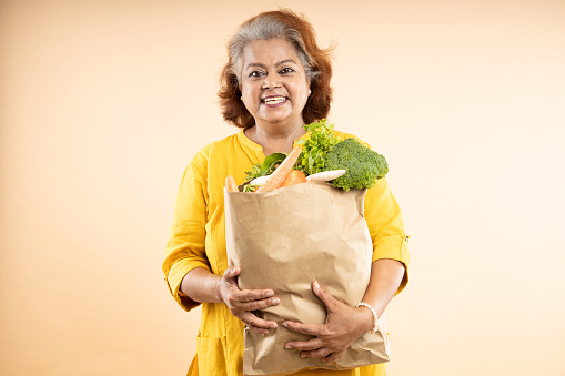 Happy senior indian woman hold grocery paper bags with food isolated on beige background. studio shot.