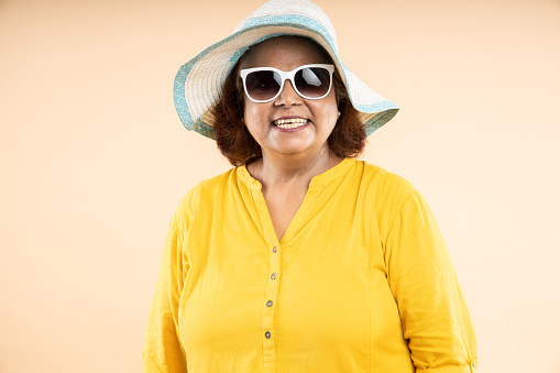 Happy senior indian woman wearing hat and sunglasses standing isolated over beige studio background, Retirement life, holidays and travel. closeup