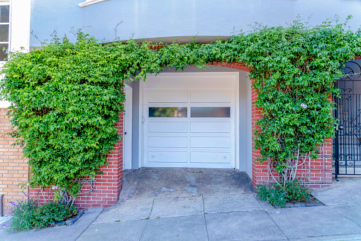 Small concrete driveway with white front door and white garage door with glass in San Francisco, CA. Garage entrance exterior with crawling shrubs against the red bricks wall at the front.