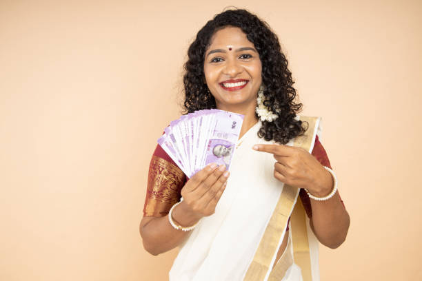Beautiful young South Indian woman wearing white saree holding money of 100 rupee notes isolated on beige background, Happy Asian female model pointing at lot of cash in hand. Beautiful young South Indian woman wearing white saree holding money of 100 rupee notes isolated on beige background, Happy Asian female model pointing at lot of cash in hand. south indian lady stock pictures, royalty-free photos & images