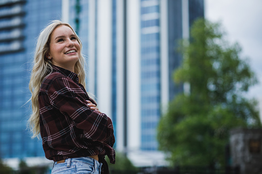 Copy space shot of joyful young blond woman standing on the city street, arms crossed, looking up, smiling and contemplating.