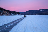 Winter travel. The car drives along an icy road along a frozen river bed against the backdrop of mountains and a sunset sky.