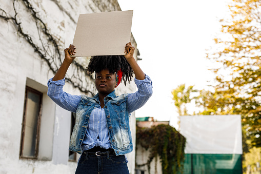 Copy space shot of confident young Black woman standing on the city street, holding a cardboard cut out and protesting for a social cause.