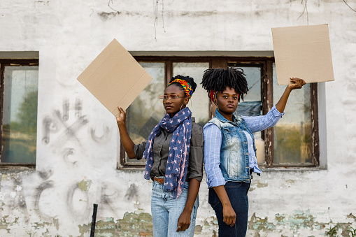 Portrait of two confident young Black female activists standing back to back, holding cardboard cut outs, with copy space, and protesting on the city street.