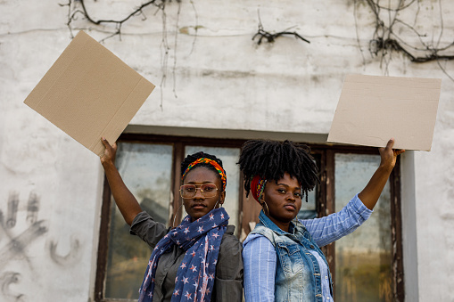 Portrait of two young Black female activists standing back to back, holding cardboard cut outs, with copy space, and protesting on the city street. One of them is looking at camera with confidence.