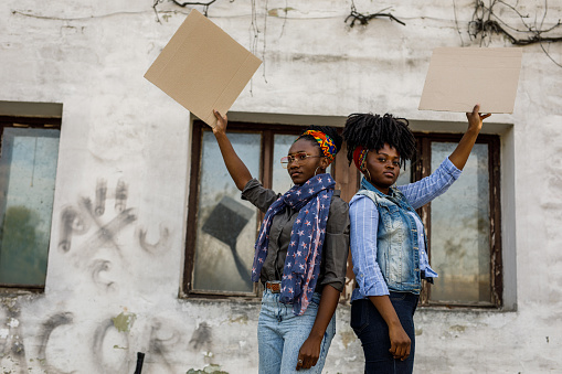 Portrait of two serious young Black female activists standing back to back, holding cardboard cut outs, with copy space, and protesting on the city street. One of them is looking at camera with confidence.