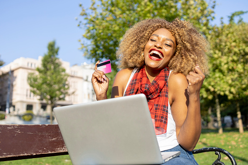 Extremely happy young woman using laptop and credit card at the park