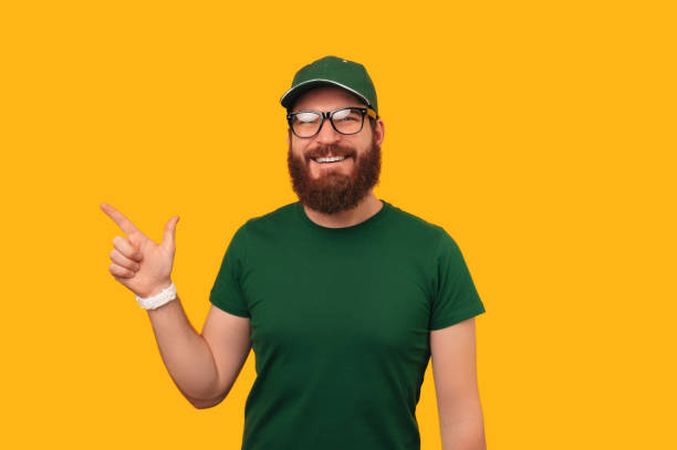 istock Young bearded delivery man wearing green uniform and cap is pointing aside. 1447882770