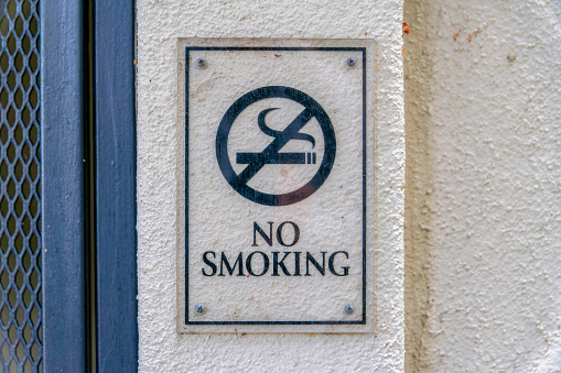 Acrylic no smoking signage against the beige wall in Silicon Valley, San Jose, California. Close-up no smoking sign with symbol near the dark gray metal mesh wall on the left.