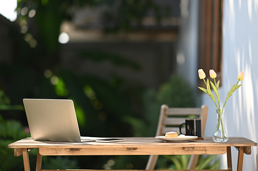 Laptop computer, cup of coffee, notebook and flower pot on wooden table at outdoor.