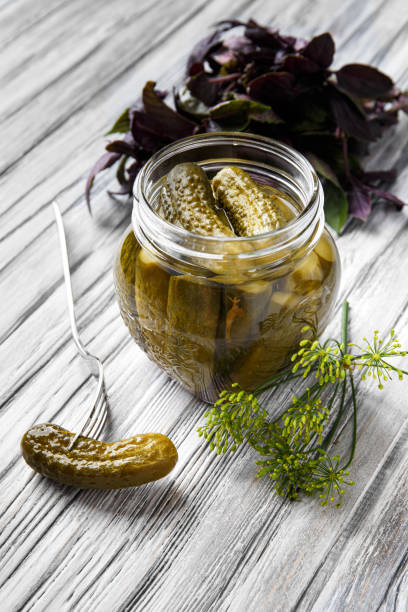 istock Open jar with pickled cucumbers and cucumber on a fork on a light wooden background 1447889514