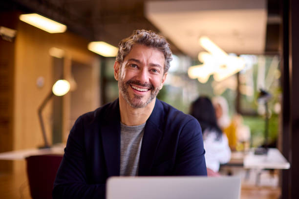 porträt eines reifen geschäftsmannes, der am schreibtisch im büro am laptop arbeitet - business businessman cheerful happiness stock-fotos und bilder
