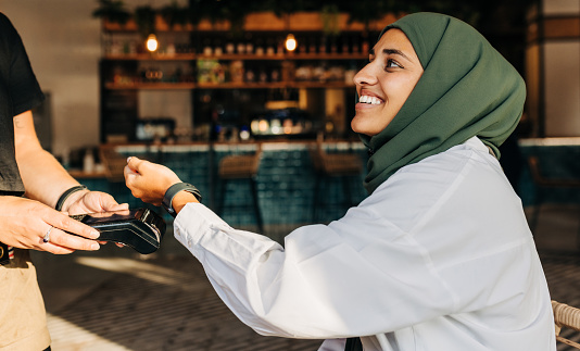 Happy Muslim woman scanning her smartwatch on a card machine to pay her bill in a cafe. Woman with a hijab smiling cheerfully while doing a contactless transaction using NFC technology.
