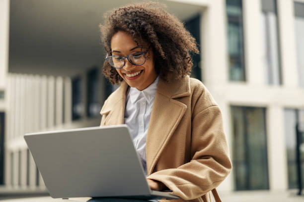 Happy african business woman working laptop while sitting outside on background of office building stock photo