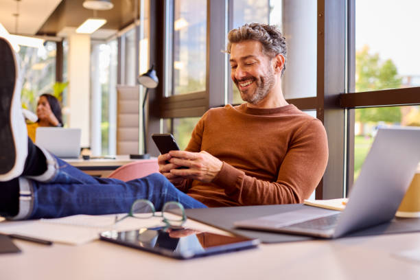 Casually Dressed Mature Businessman With Feet On Desk In Office Using Mobile Phone stock photo