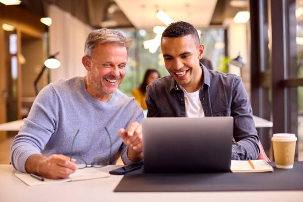 机の上でノートパソコンで作業する若い同僚を指導する成熟した実業家 - learning male studying smiling ストックフォトと画像