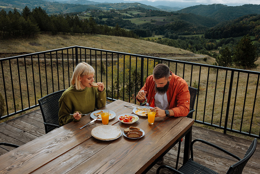 A young beautiful Caucasian couple is having breakfast on the patio of their mountain cabin .