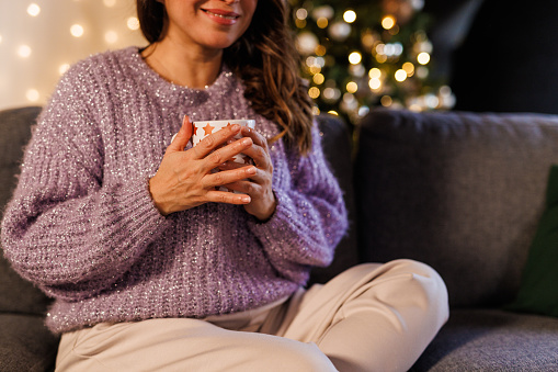 Portrait of happy mid adult woman sitting on the sofa in her living room, enjoying a warm cup of tea, looking away and contemplating about Christmas holidays. Glistening Christmas tree in the background.
