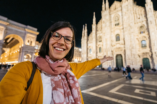 Beautiful young female tourist taking a selfie in front of the cathedral in Milan, Italy at night.