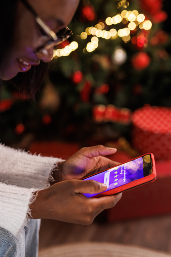 Close up shot of young Black woman sitting by the Christmas tree and rating her experience after doing online shopping via smart phone.