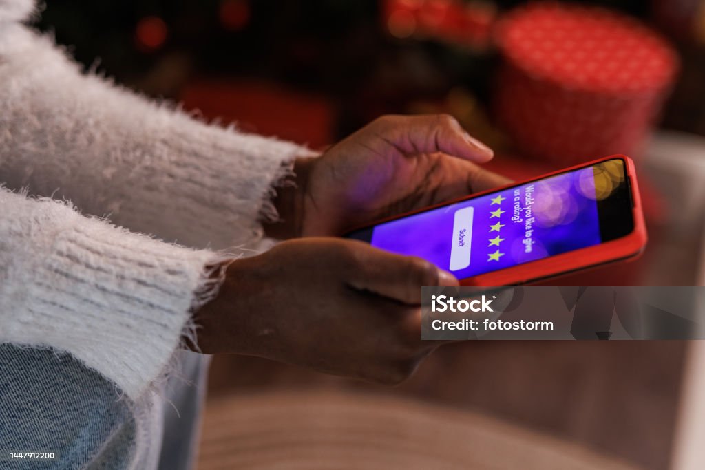 Cut out shot of woman doing online Christmas shopping via smart phone Close up shot of unrecognizable young Black woman sitting by the Christmas tree and rating her experience after doing online shopping via smart phone. Rating Stock Photo