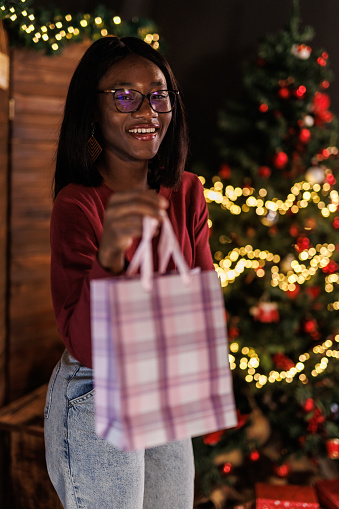 Portrait of joyful young woman standing by the Christmas tree and smiling at camera while giving you a gift bag.