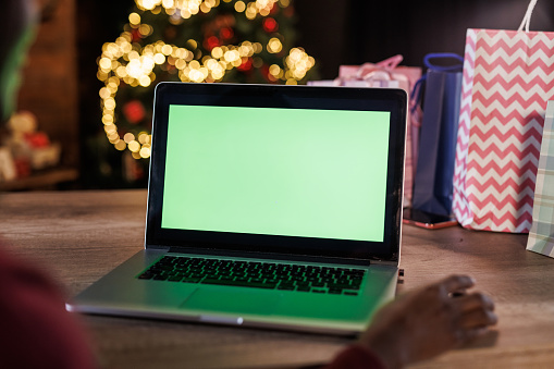 Close up shot of young Black woman sitting at her home office desk and using laptop with chroma key on the screen. Glistening Christmas tree in the background.