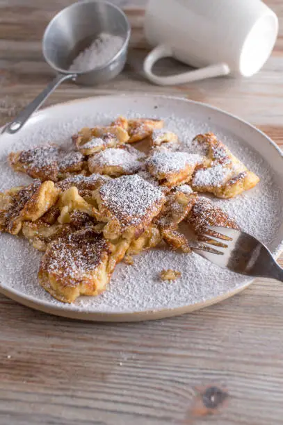 Traditional homemade kaiserschmarrn, austrian pancake with powdered sugar topping. Served on a white plate on light wooden background with kitchen utensils. Closeup with copy space