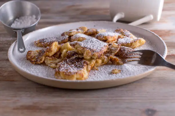 Homemade fresh baked austrian shredded pancake, called kaiserschmarrn. Topped with powdered sugar and served ready to eat on a white plate on wooden table. Closeup, front view with copy space
