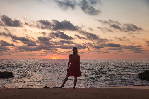 Rear view silhouette young woman standing on sandy beach at evening sea background at sunset. From behind slim lady wear red dress on tropical dusk ocean. Travel vacation concept. Copy text space