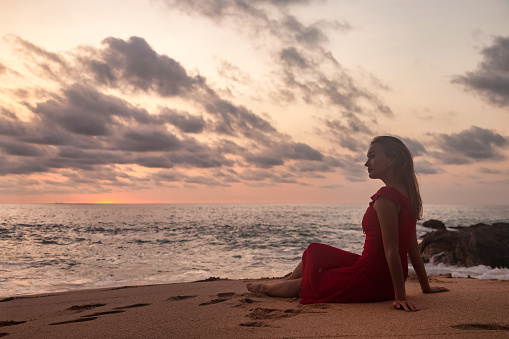 Alone young woman wear red dress sitting on sandy beach at evening sea background, thinking looking away. Lonely pretty lady on tropical ocean at sunset. Travel vacation concept. Copy text space