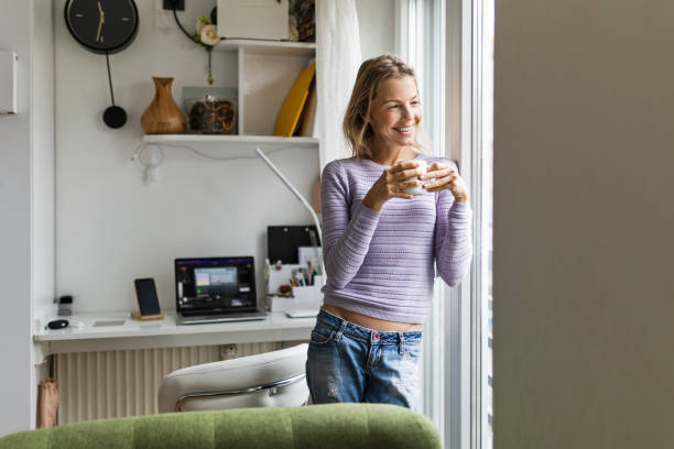 istock Smiling young woman with coffee looking through the window 1447940104