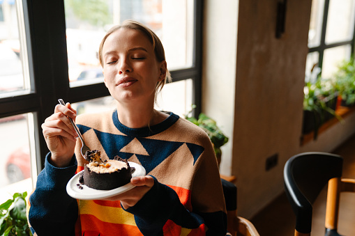 Young woman in colorful sweater eating delicious dessert while sitting at table in cafe