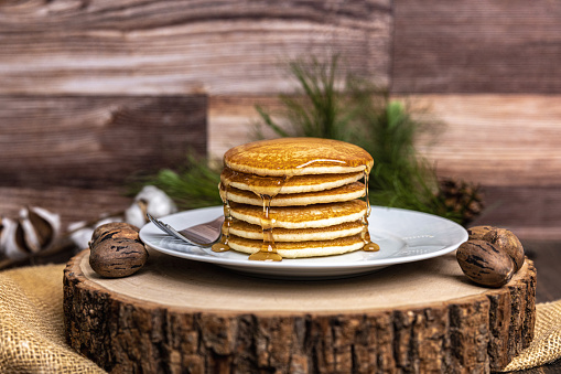 A large plate stack of pancakes with a fork against a wood background plain with syrup, pecans and a rustic background with southern cotton and pine decor.