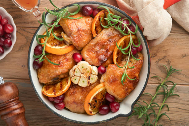 gebackene hähnchenschenkel mit orange, cranberry und würzigen kräutern rosmarin in der pfanne auf hellem rustikalem holzhintergrund. festliches weihnachtsabendessen-konzeptmenü. draufsicht. speicherplatz kopieren - chicken thighs stock-fotos und bilder