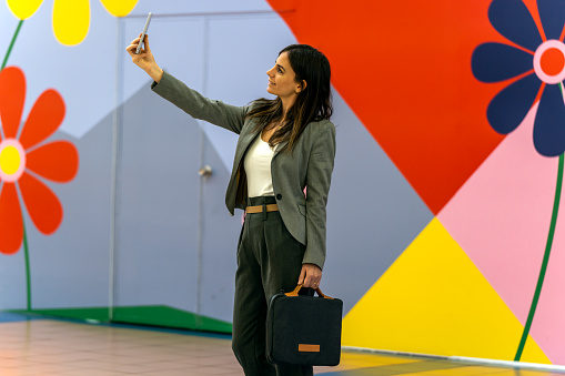 Female entrepreneur in smart casual clothes with briefcase smiling and taking selfie against colorful wall with geometric ornament and flowers