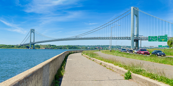 High resolution stitched panorama of the Verrazano-Narrows Bridge on a spring morning with Cars driving on the Belt Parkway. The bridge connects boroughs of Brooklyn and Staten Island in New York City. The bridge was built in 1964 and is the largest suspension bridge in the USA. Historic Fort Wadsworth is under the bridge. The photo was taken from the Shore promenade in Bay Ridge Brooklyn. Canon EOS 6D full frame censor camera. Canon EF 85mm f/1.8 USM Prime Lens. 2:1 Image Aspect Ratio. This image was downsized to 50MP. Original image resolution was 68.3MP or 11686 x 5843 px.