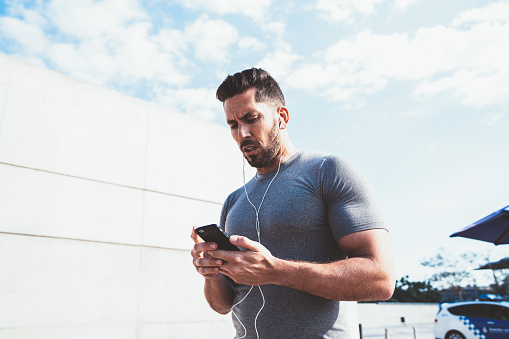 Serious caucasian male jogger using smartphone application for trekking distance of cardio workout standing outdoors, confident sportsman downloading music to mobile phone player for training