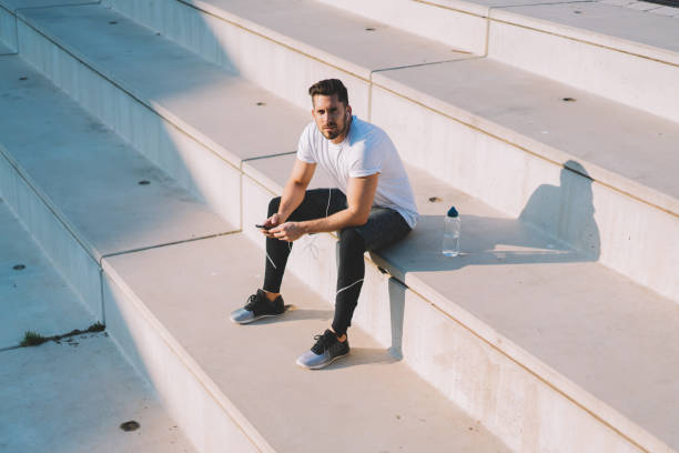istock Handsome muscular hipster guy in active clothes having rest after workout on stadium looking at camera, young 20s male using smartphone and earphones for listening motivating music on trainings 1447958651