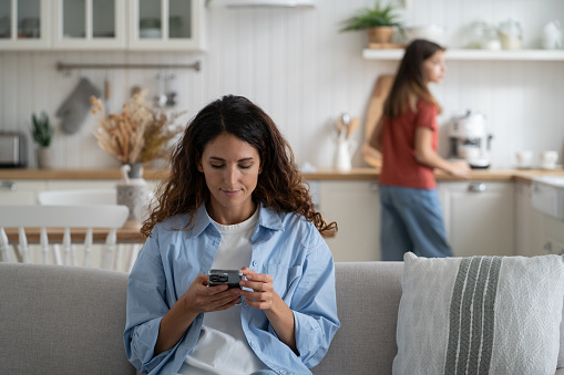 Young stay-at-home mother holding smartphone in hands, relaxing on sofa at home while her daughter cooking alone in kitchen. Woman housewife ordering food with phone, buying groceries online