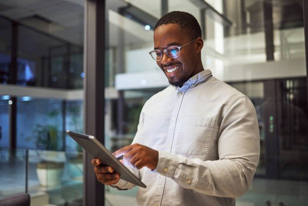 affari notturni. tablet e uomo nero che digitano e leggono informazioni online facendo ricerche e sorridendo ai buoni risultati lavorando fino a tardi. maschio felice su internet per la strategia di marketing nell'ufficio della nigeria - one young man only immagine foto e immagini stock