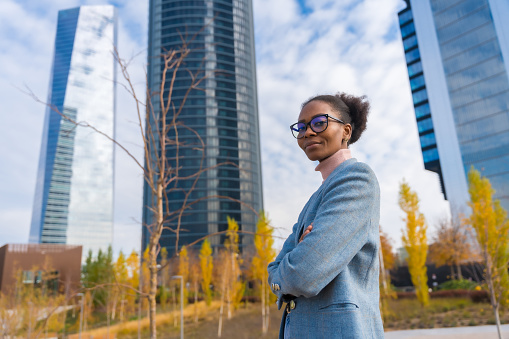 Black ethnic businesswoman or executive wearing glasses standing in a business park, arms crossed