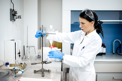 Female scientist with brown hair seen wearing blue gloves and protective eyeglasses while working on an experiment in a laboratory using test tubes during some research.