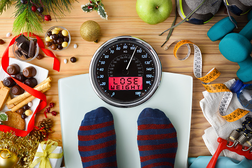 Person weighing on a scale after the Christmas holidays with message lose weight on wooden background with sweets and sports equipment. Top view.