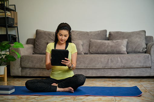 Asian Female yoga instructor sitting on yoga mat teaching input data on digital tablet at home.