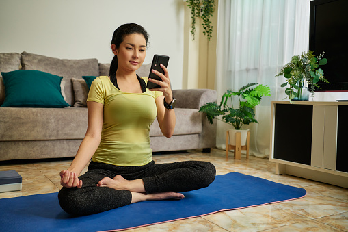 Asian Female yoga instructor sitting on yoga mat  input data on Smart phone at home.