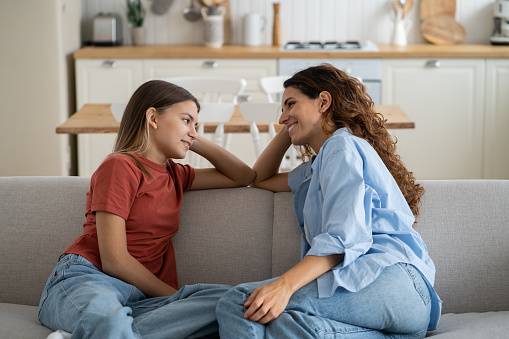 Loving mother listening to daughter with empathy and understanding while sitting together on sofa bonding at home, pre-teen girl child sharing secrets with mom, parent communicating with teenager